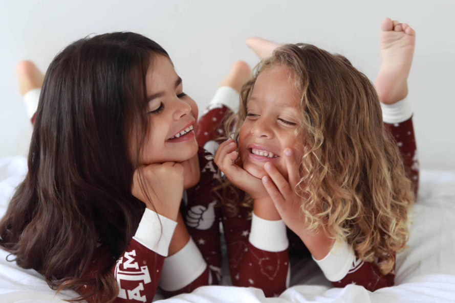 Two children in matching Texas A&M bamboo pajamas sitting on a bed, laughing and smiling.